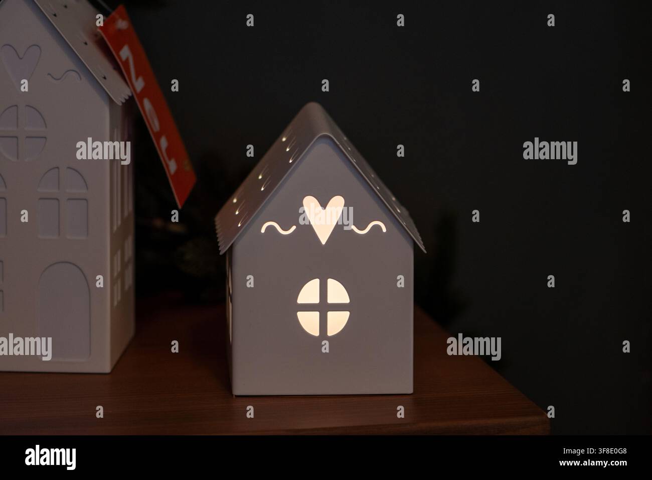 Una piccola lanterna a forma di casa bianca con ritagli a forma di cuore e finestra brilla su una superficie di legno contro uno sfondo scuro. Foto Stock