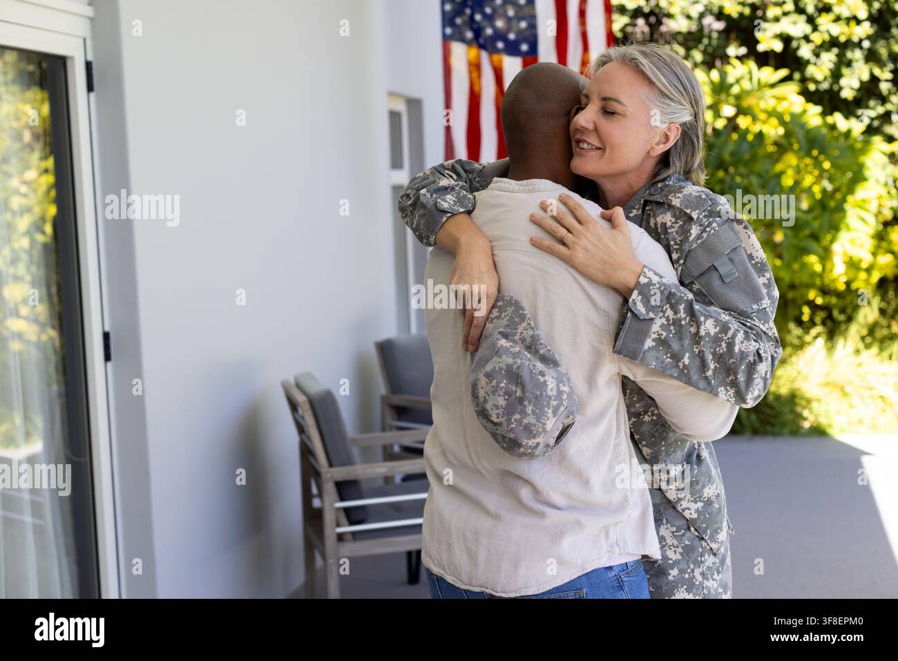 Coppia diversa che si abbraccia sul portico anteriore, donna in mimetica che tiene un berretto da pattuglia vicino alla bandiera americana Foto Stock