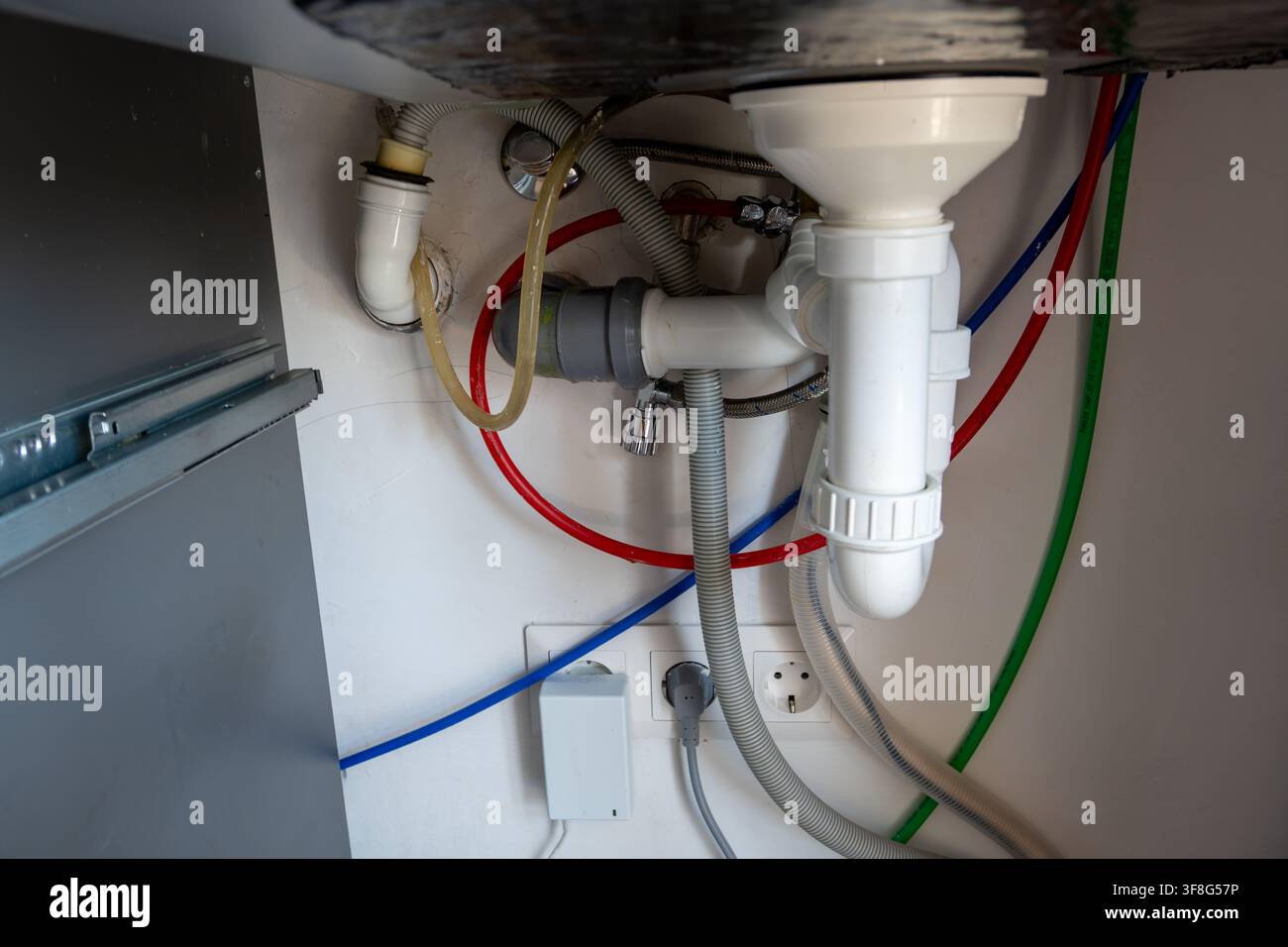 Rete complessa di tubi di scarico in plastica, tubi flessibili e linee di approvvigionamento idrico colorate installate sotto il lavandino domestico con prese di corrente visibili. Foto Stock