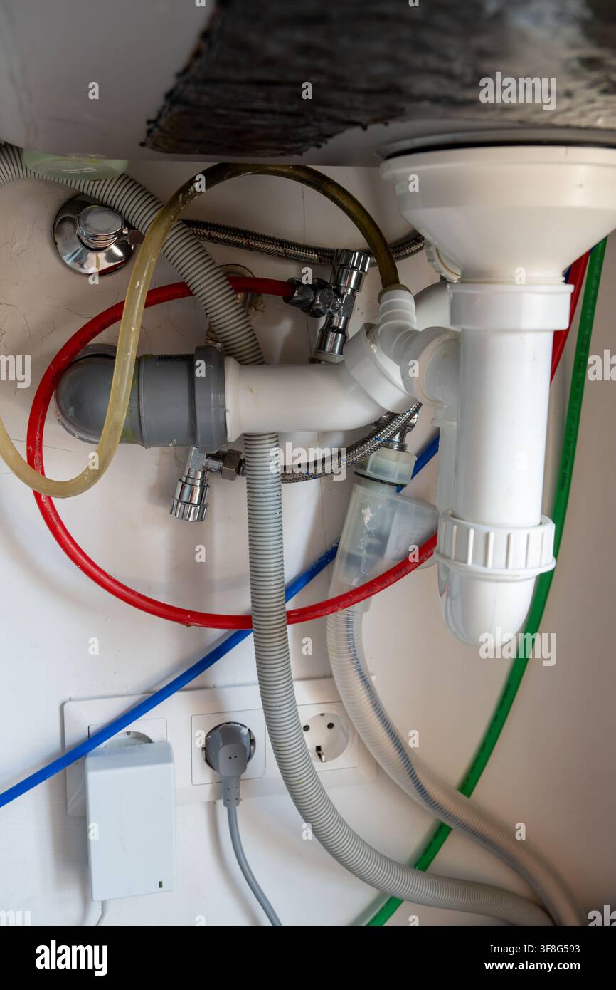 Vista dettagliata dei collegamenti idraulici domestici con sifone bianco, tubi di scarico grigi, condotte dell'acqua colorate e prese elettriche a muro sotto la cucina Foto Stock