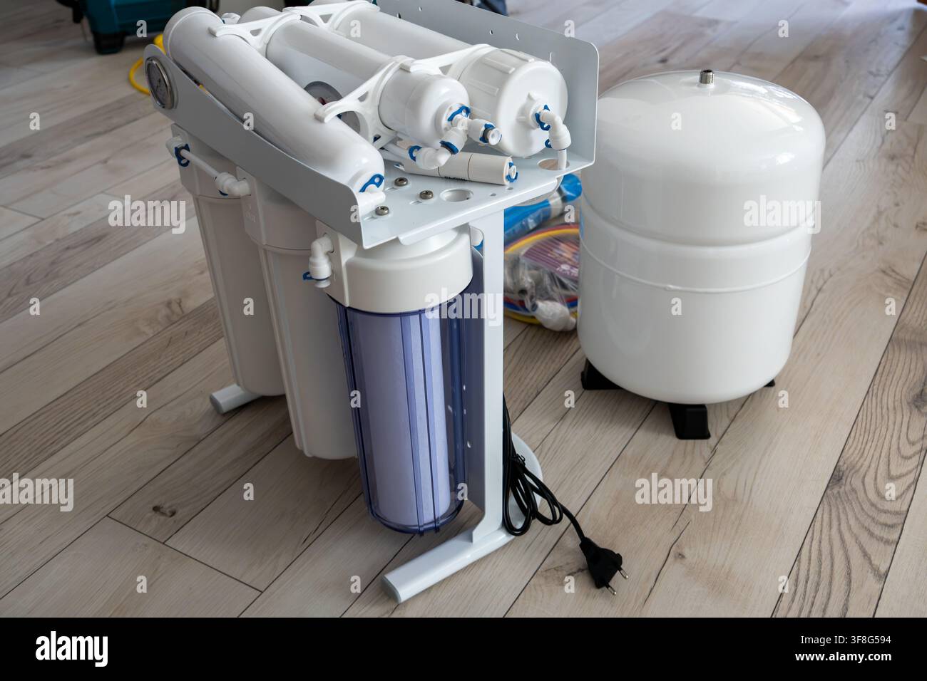 Moderno sistema di filtro per l'acqua ad osmosi inversa con cartucce multisterreno e serbatoio bianco posizionato su pavimento di legno, preparato per l'installazione domestica Foto Stock