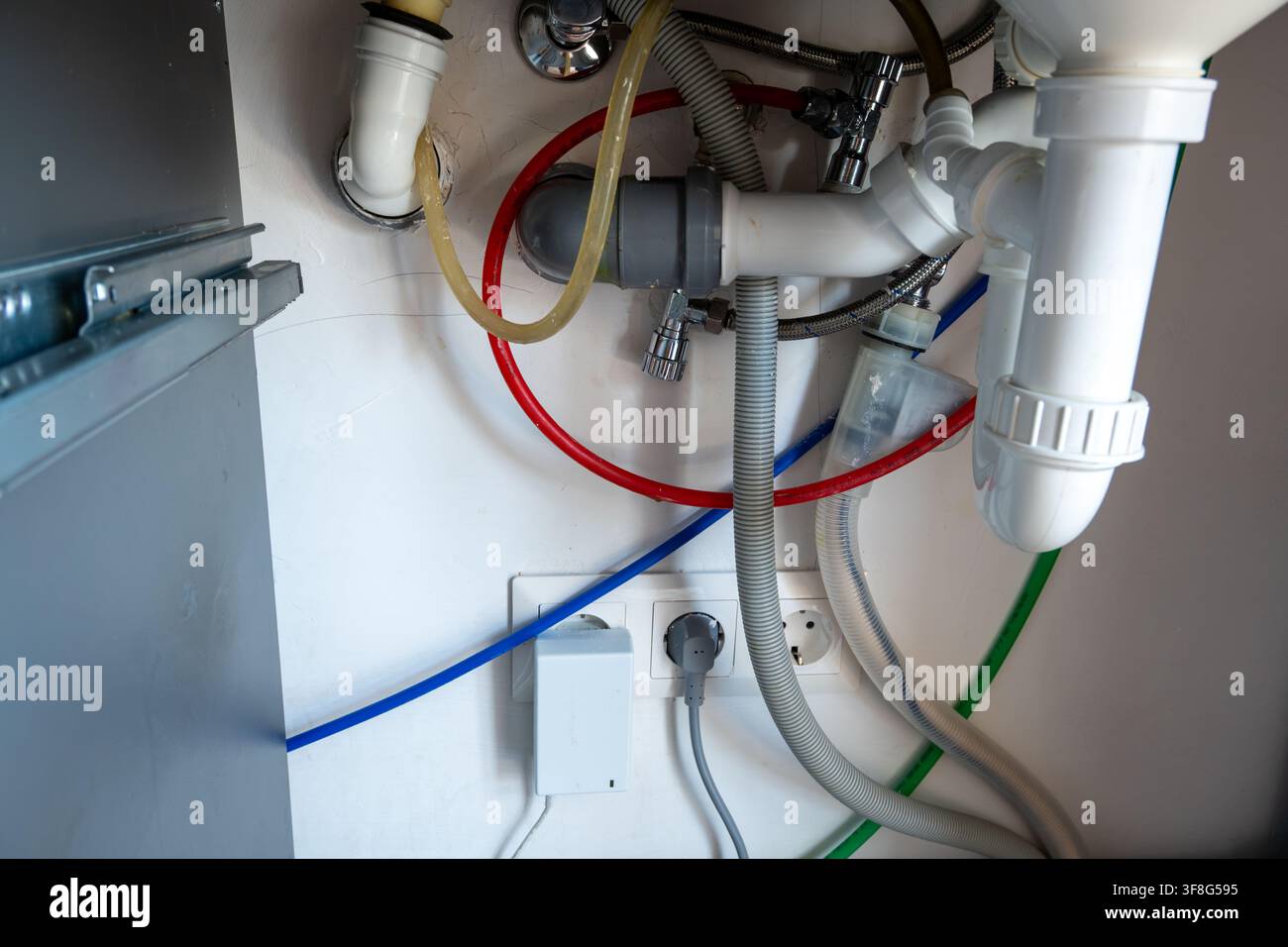 Primo piano di un complesso sistema idraulico per lavello da cucina con tubi di plastica, tubi flessibili di scarico, condotte dell'acqua colorate e prese elettriche a muro. Foto Stock