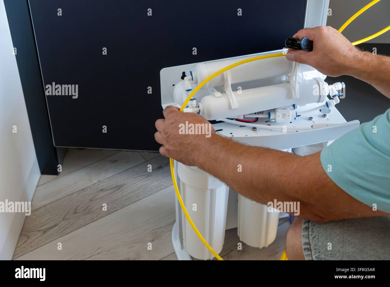 Primo piano di mani maschili che collegano un tubo di plastica giallo a un sistema di purificazione dell'acqua ad osmosi inversa durante l'installazione di un filtro domestico su un pavimento di legno Foto Stock