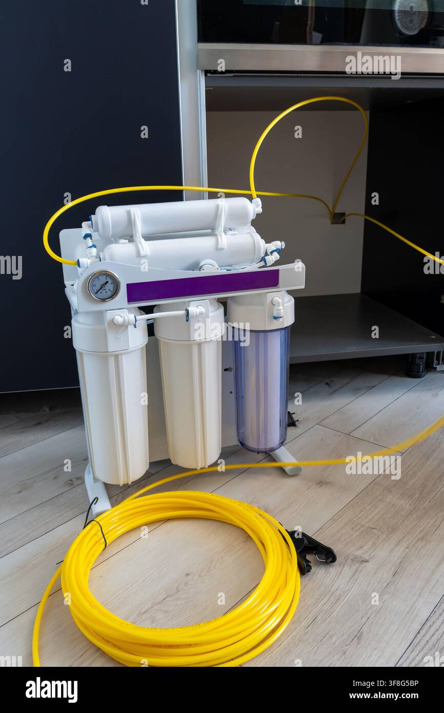 Sistema di purificazione dell'acqua a osmosi inversa multistadio con manometro e bobina di tubo di plastica giallo si trova su pavimento di legno accanto alla cabina della cucina Foto Stock