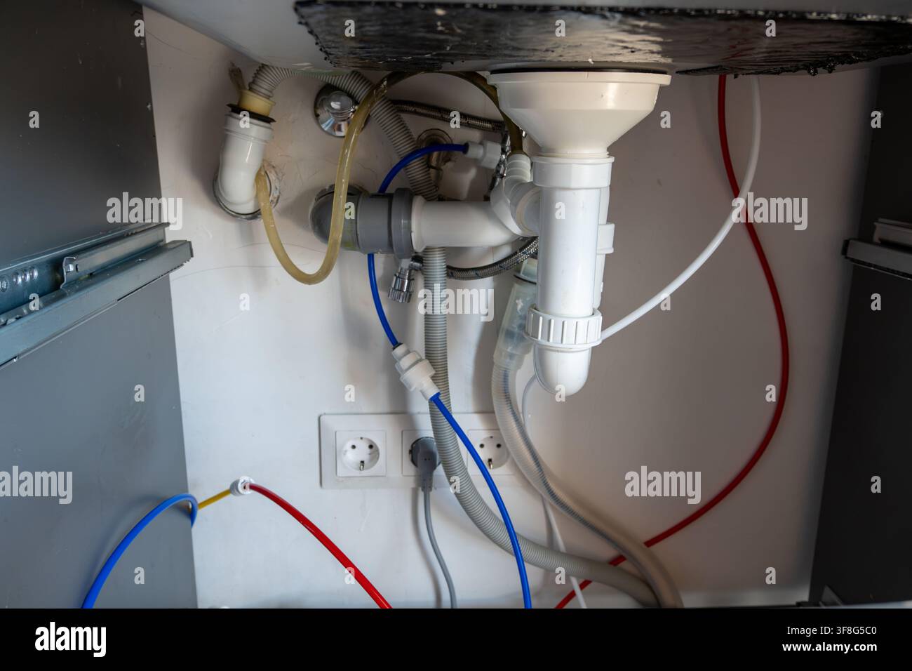 Vista sotto il lavandino di un mobiletto idraulico da cucina con tubi di connessione per sifone di scarico bianchi, tubi flessibili, prese elettriche e nuovo contenitore filtro colorato Foto Stock