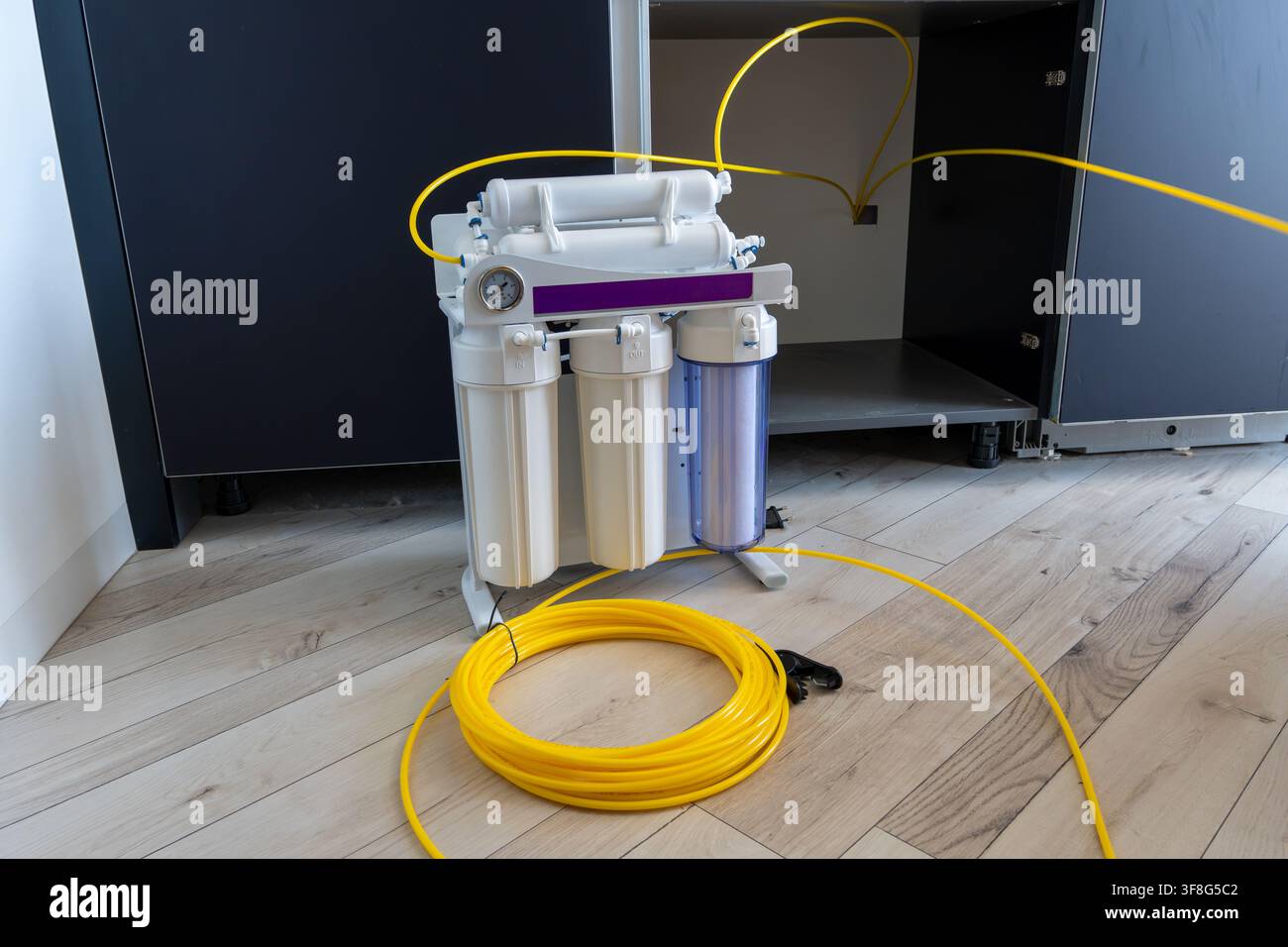 Unità di filtro per osmosi inversa con tubi gialli collegati e bobina di tubo in plastica posizionata su pavimento in legno davanti al mobiletto aperto sotto il lavello in cucina Foto Stock