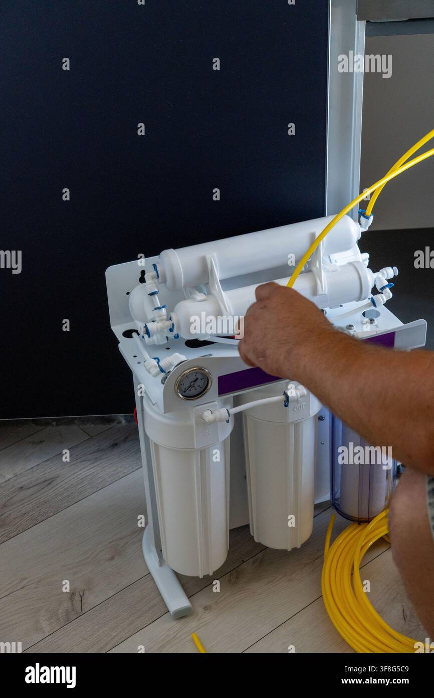 Mano di uomo che inserisce un tubo di plastica giallo in un sistema di filtro per osmosi inversa durante il processo di installazione domestica su uno sfondo di pavimento in legno. Foto Stock