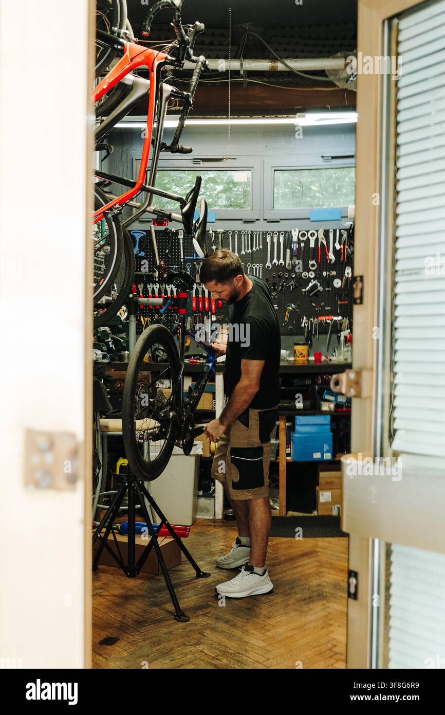 Meccanico ciclista maschile effettua la manutenzione di una bicicletta su un banco di riparazione all'interno di un'officina compatta. La vista attraverso la porta mostra un servizio professionale per biciclette Foto Stock