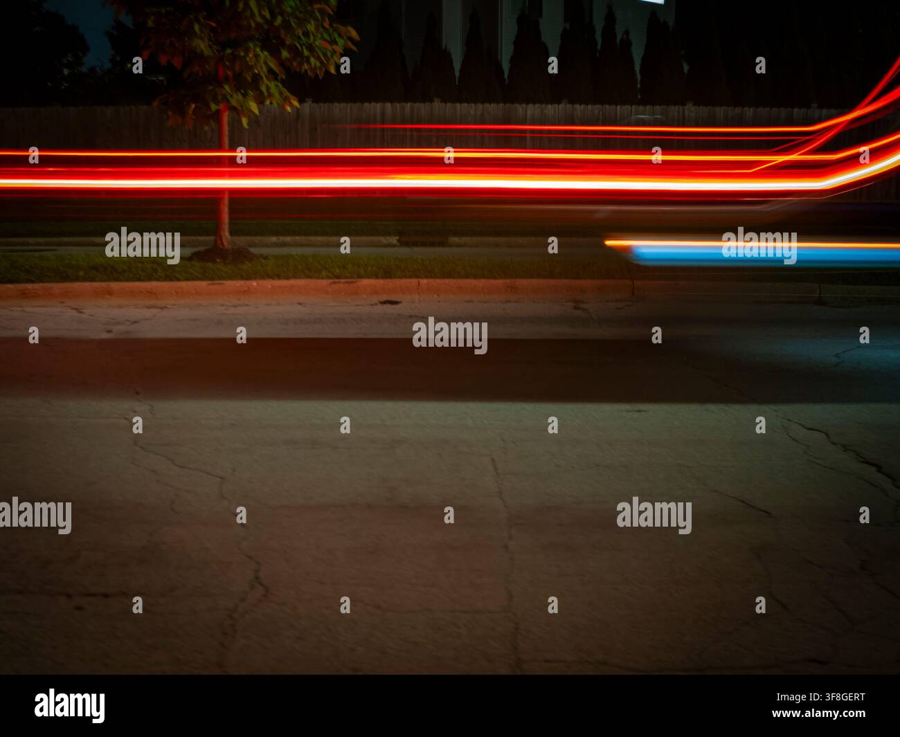Fotografia notturna a lunga esposizione che mostra vivaci tracce di luce rosse e blu provenienti da veicoli in movimento che sfrecciano lungo una strada di periferia e alberi Foto Stock