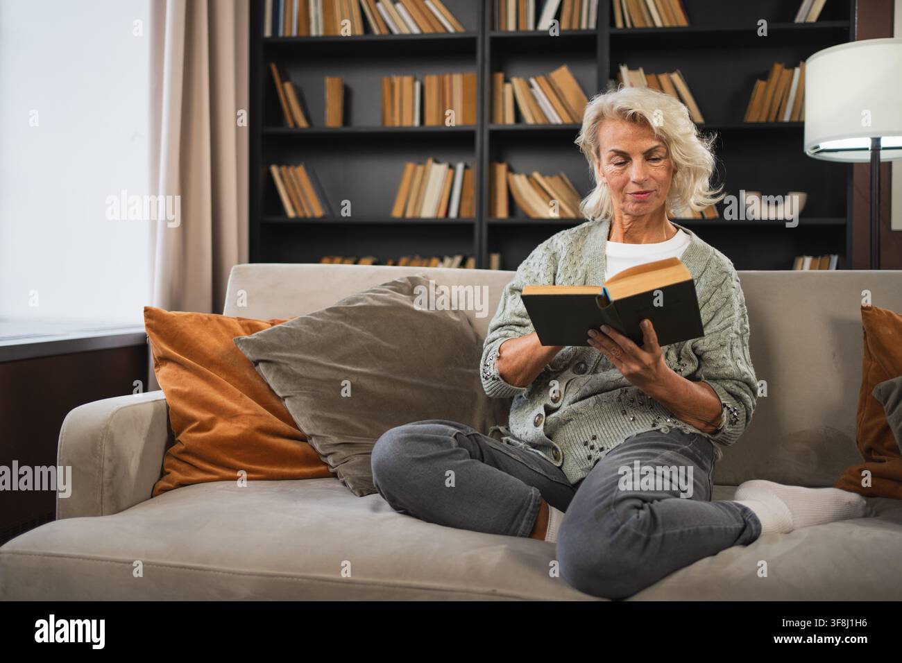 Comfort rilassato anziano anziano anziano anziano donna che legge un libro a casa. Donna matura che riposa goditi una giornata tranquilla seduto sul divano di casa da sola. Donna adulta che passa il tempo libero a tenere i libri sorridenti. Concetto di pensione Foto Stock