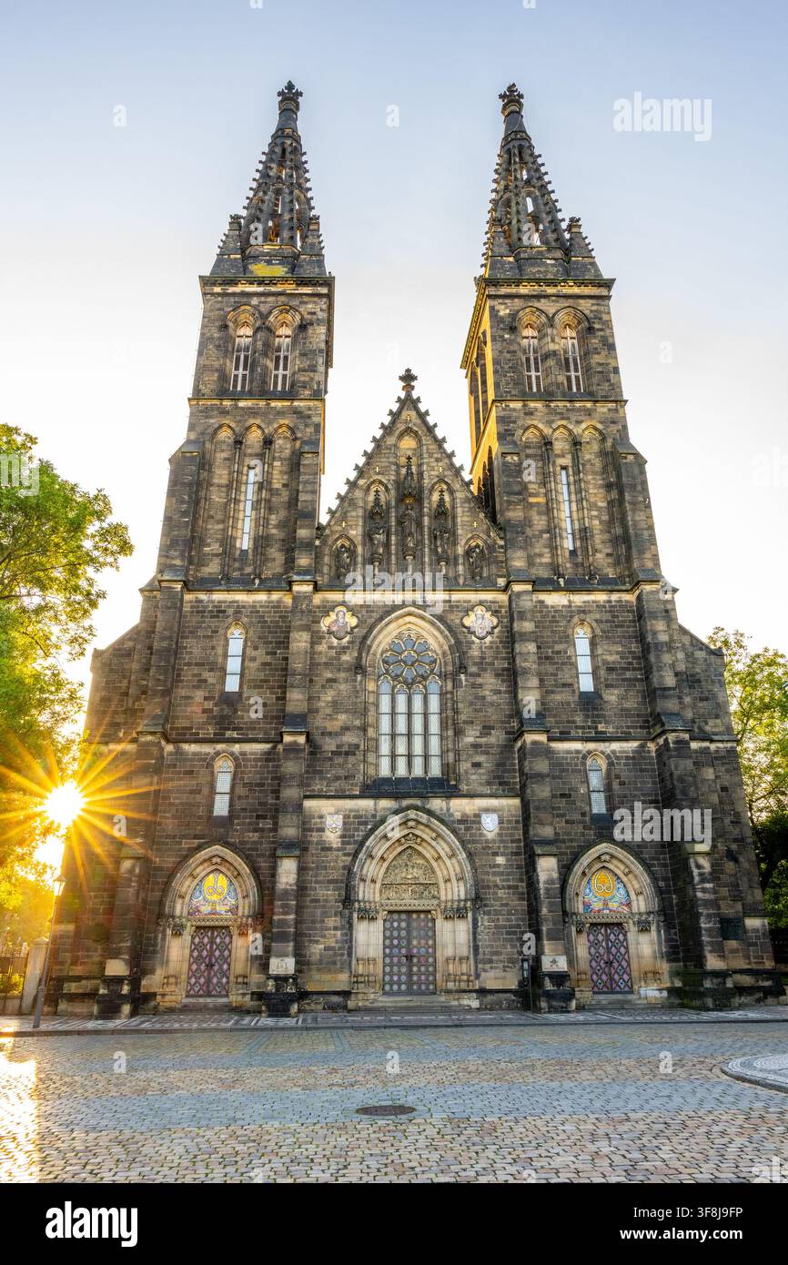 La Basilica di San Pietro e San Paolo si erge imponente a Vysehrad. La luce del sole splende da dietro la chiesa, evidenziando le sue torri e i dettagli in pietra. L'area è tranquilla e aperta. Foto Stock
