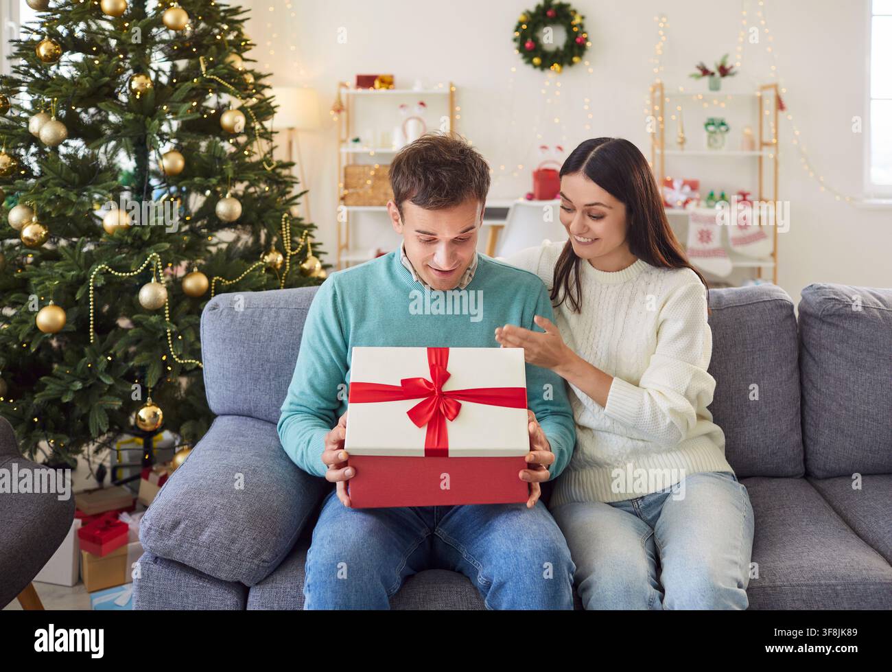 Giovane uomo apre con entusiasmo un regalo di Natale e guarda all'interno della scatola con sincera ammirazione. Foto Stock