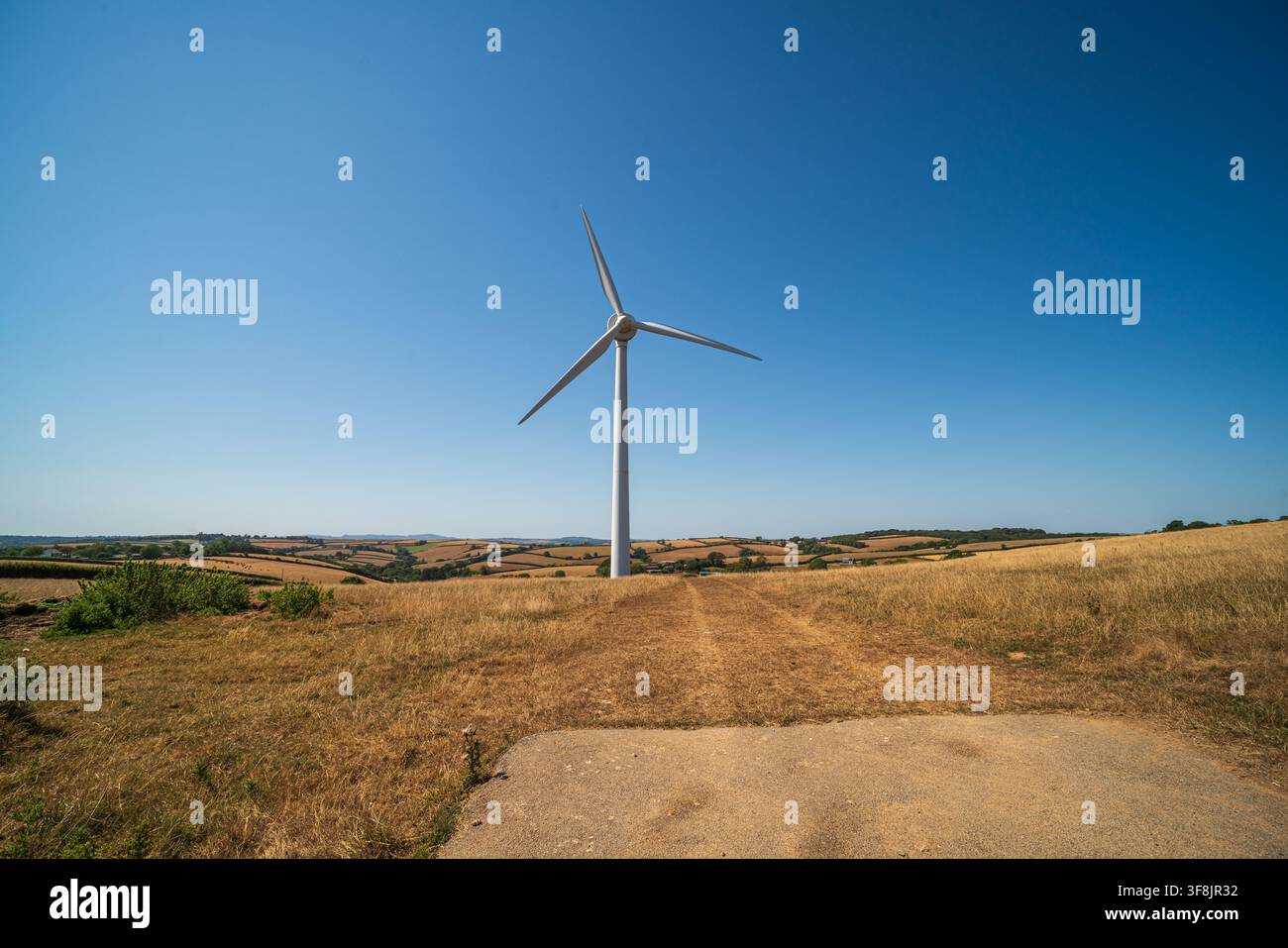 Una moderna turbina eolica si erge alta su una collina erbosa arida sotto un cielo blu chiaro e luminoso, affacciandosi sui campi agricoli a chiazze del Cornwall. Foto Stock