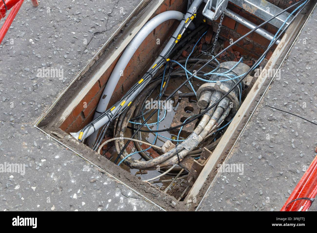 Bobine di cavo di traino grigio e cavo a fibre ottiche nere riposano con una guida per cavi gialla accanto a una barriera rossa e bianca in un cantiere stradale. Il tiro Foto Stock