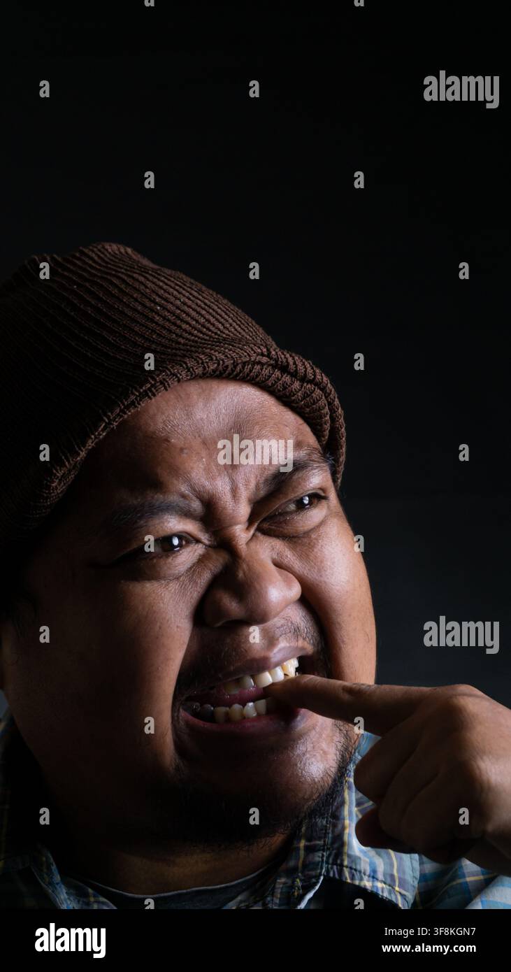 Un ritratto in primo piano di un giovane uomo asiatico dell'Indonesia del Sud-est che si pulisce i denti con un dito mostrando un'espressione facciale smorfiosa contro un Da Foto Stock