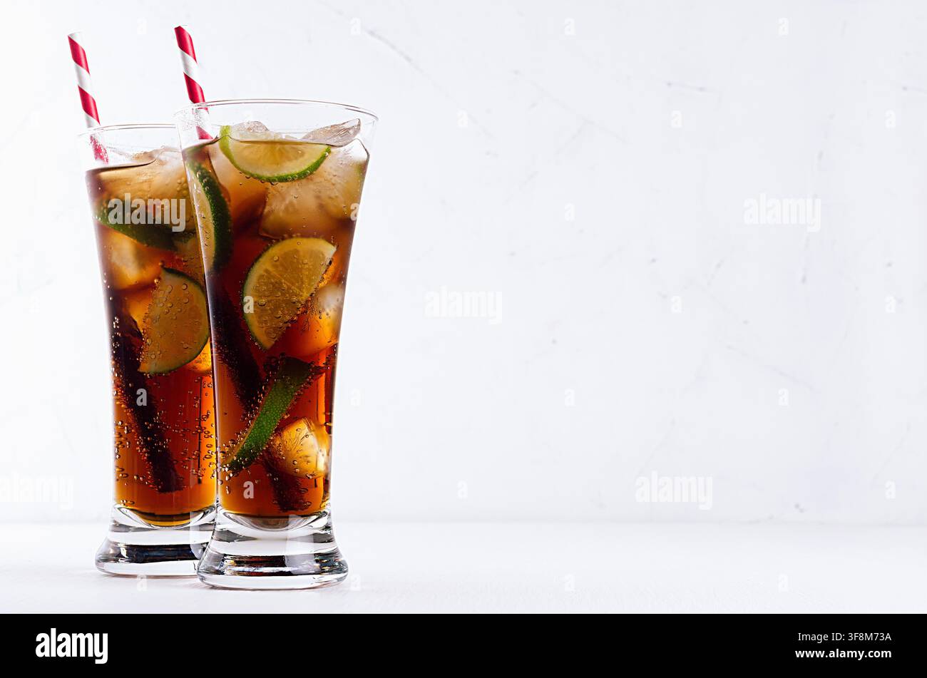 Cocktail freddo cuba libre in due lunghi bicchieri con paglia, pezzi di lime su morbido sfondo bianco moderno. Foto Stock