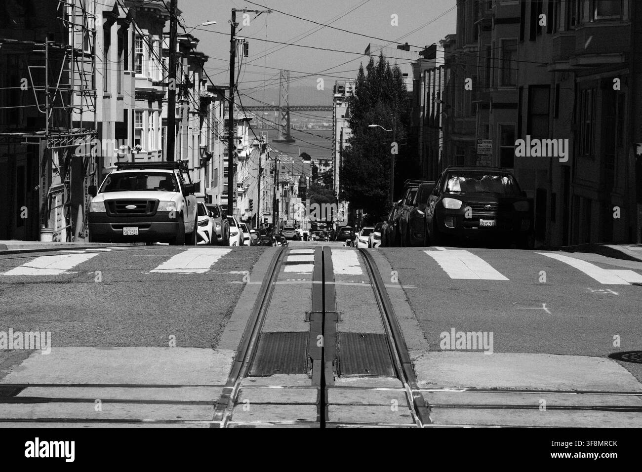 I binari del teleferico scendono una strada ripida verso la baia di San Francisco, in California, con veicoli parcheggiati e edifici urbani che costeggiano la strada. Foto Stock