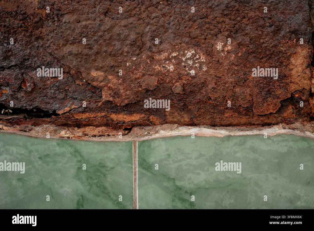 Bordo metallico scuro pesantemente arrugginito che incontra una texture superficiale di piastrelle verdi. Foto Stock