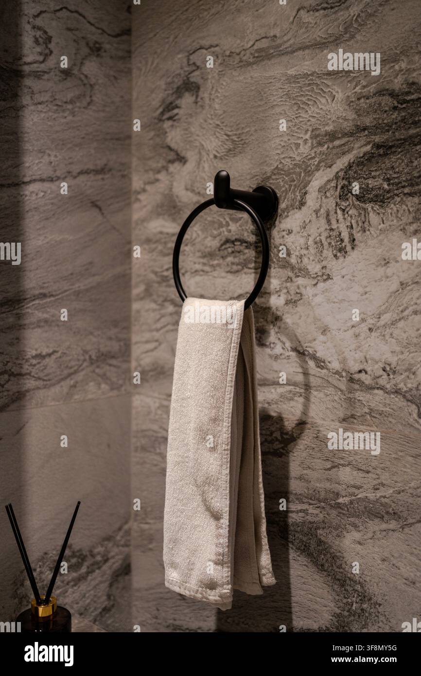 Asciugamano bianco appeso a un portaanello in metallo nero contro un muro di bagno in pietra grigia strutturata. Foto Stock