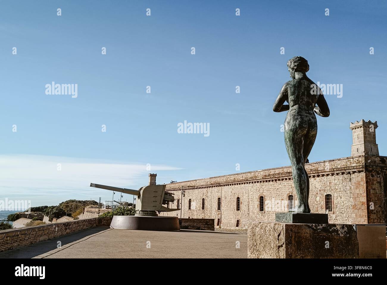 Barcellona, Spagna - 05 dicembre 2025: Vista del castello storico di Montjuïc con una statua di bronzo in primo piano e un grande cannone di difesa costiera Foto Stock