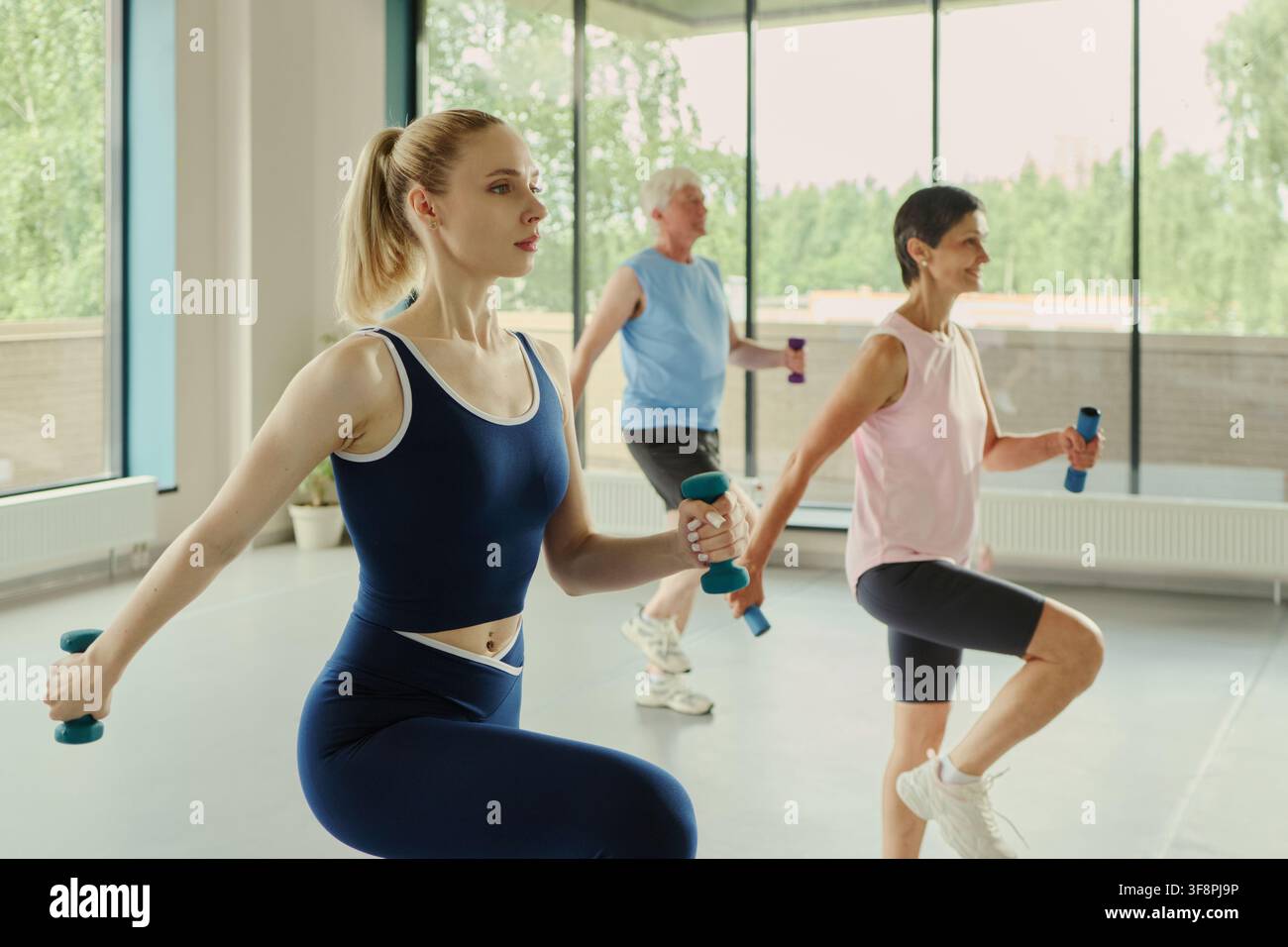 Giovane donna caucasica che conduce un corso di fitness con una donna matura e un uomo anziano che si esercitano con i manubri. L'immagine rappresenta uno stile di vita attivo e campagne di marketing del benessere Foto Stock