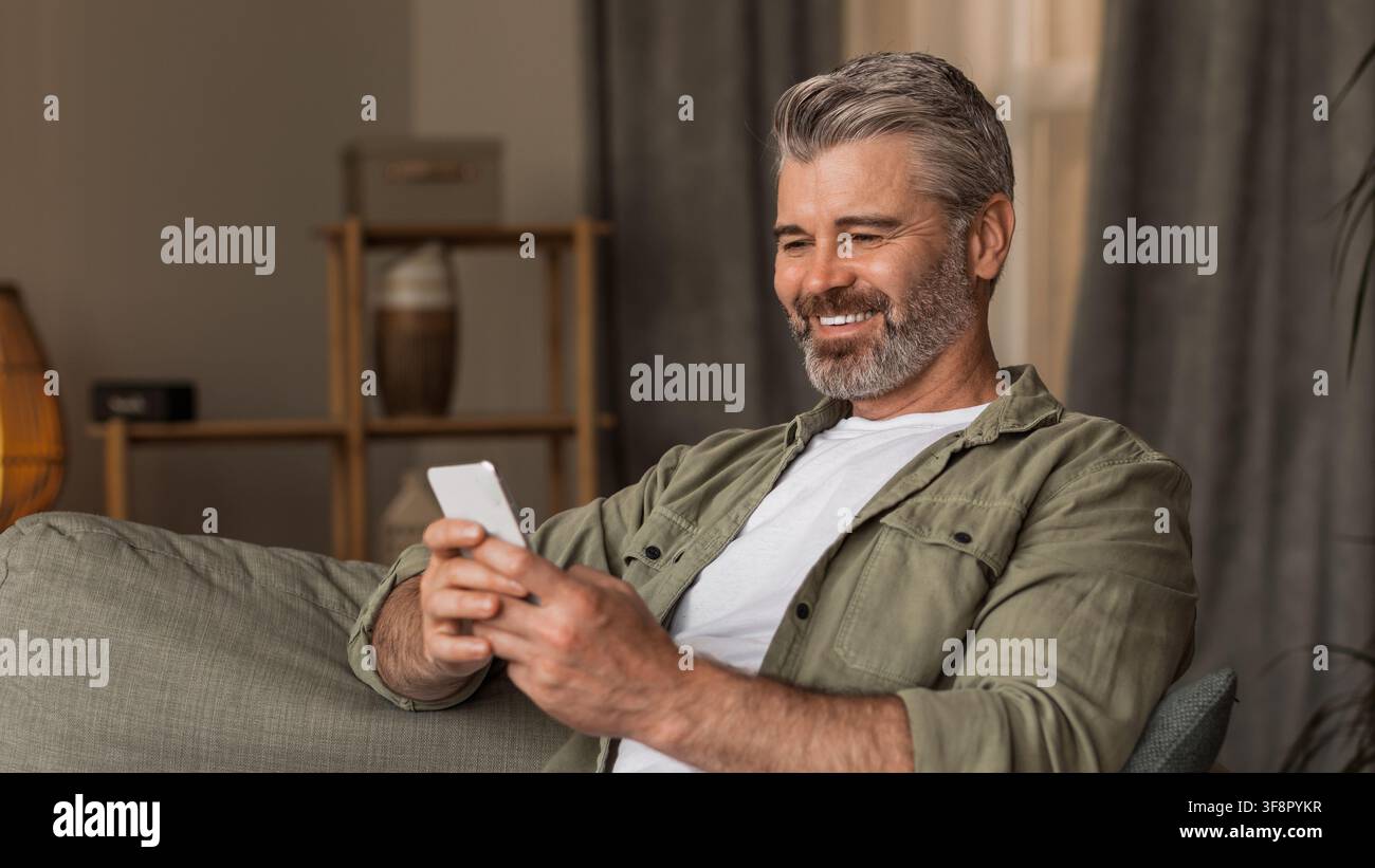 Uomo caucasico di mezza età sorridente che usa uno smartphone sul divano a casa, concetto di stile di vita rilassato Foto Stock
