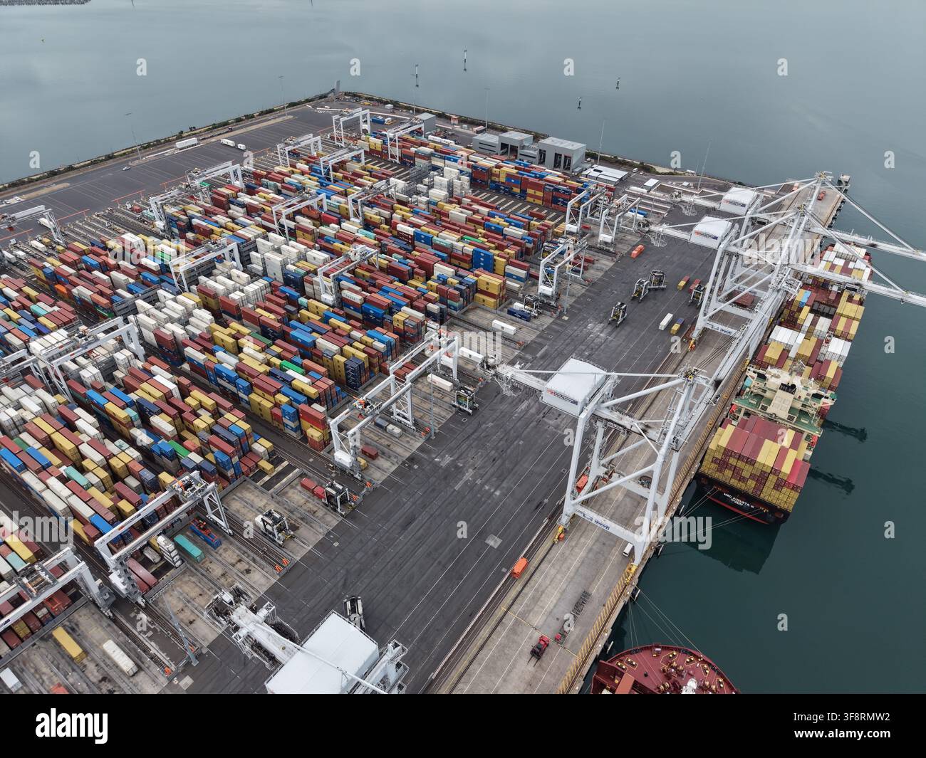 Porto commerciale affollato con container di spedizione impilati sul terminal e gru che caricano il carico su una grande nave portacontainer Foto Stock