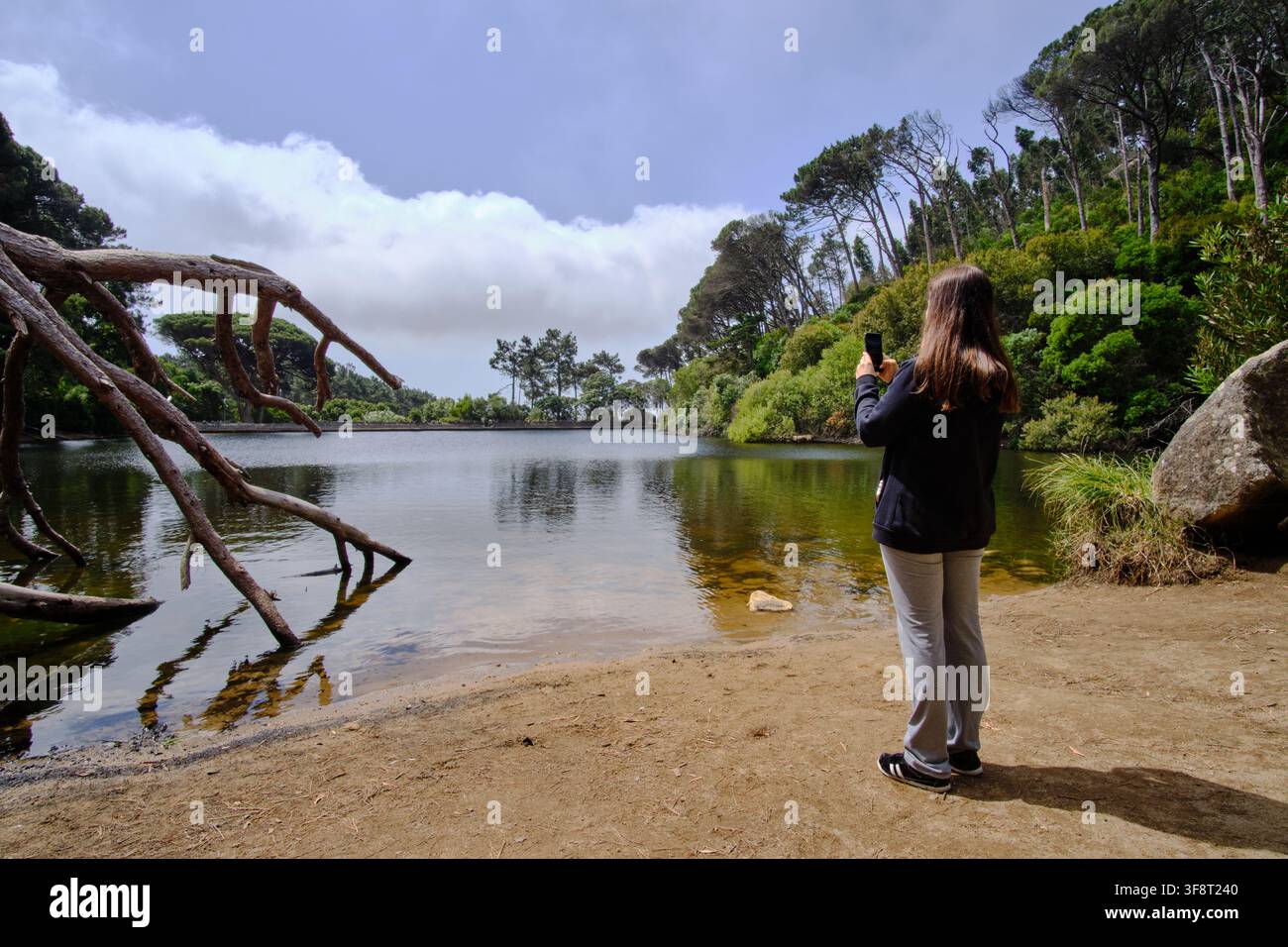 Donna in piedi vicino a un lago, che filma il paesaggio naturale con un telefono cellulare Foto Stock