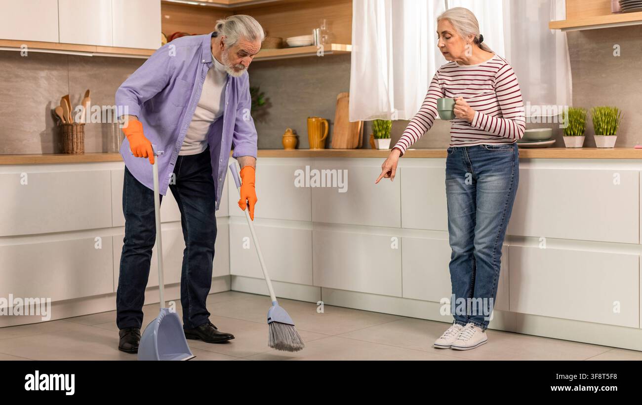 Coppia caucasica anziana che pulisce insieme la cucina, uomo che spazza il pavimento con una scopa Foto Stock