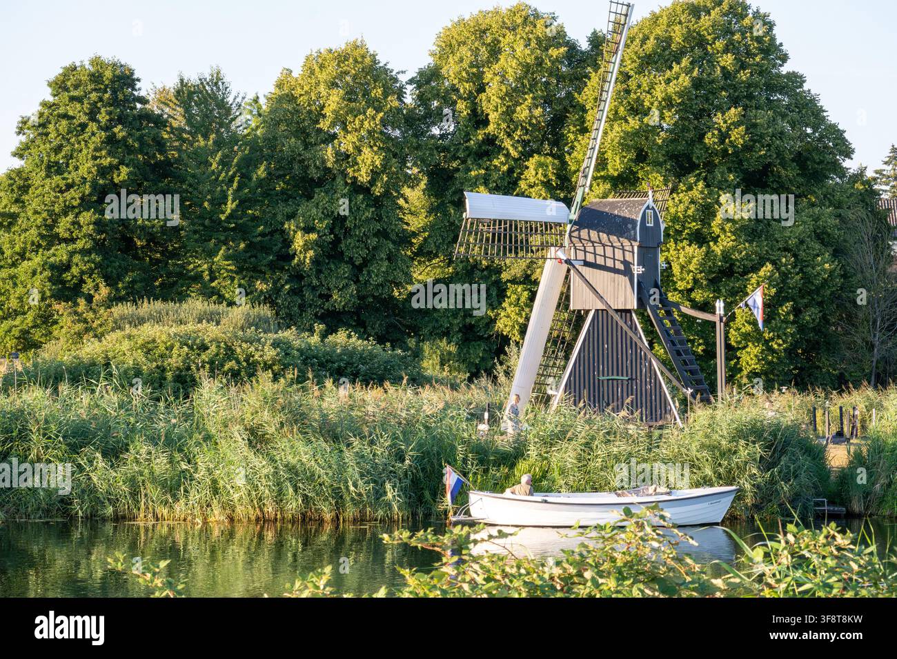 Mulino a vento tradizionale olandese in legno sulla riva del fiume con una piccola barca che naviga sul canale nella campagna di Gelderland nei Paesi Bassi Foto Stock