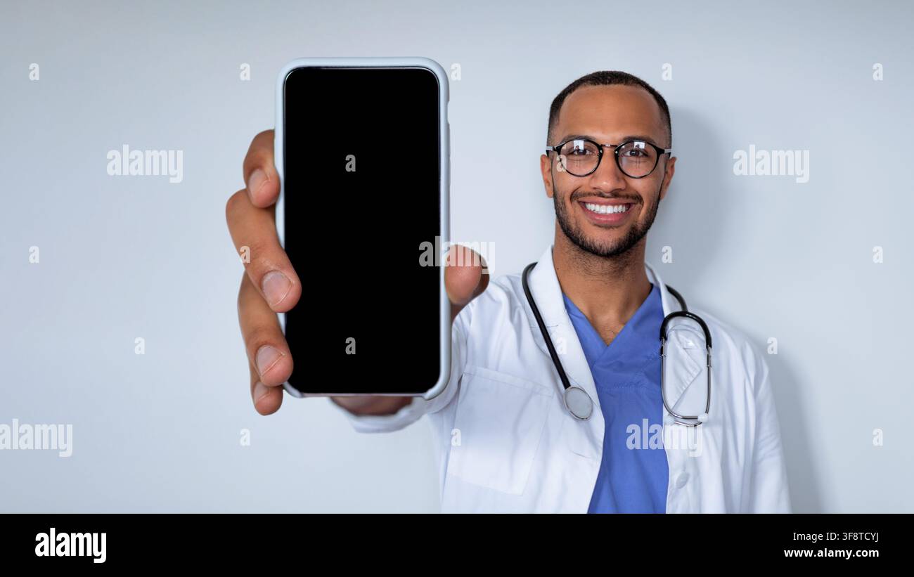 Medico nero sorridente che tiene uno smartphone con schermo vuoto, clinica. Concetto di assistenza sanitaria Foto Stock
