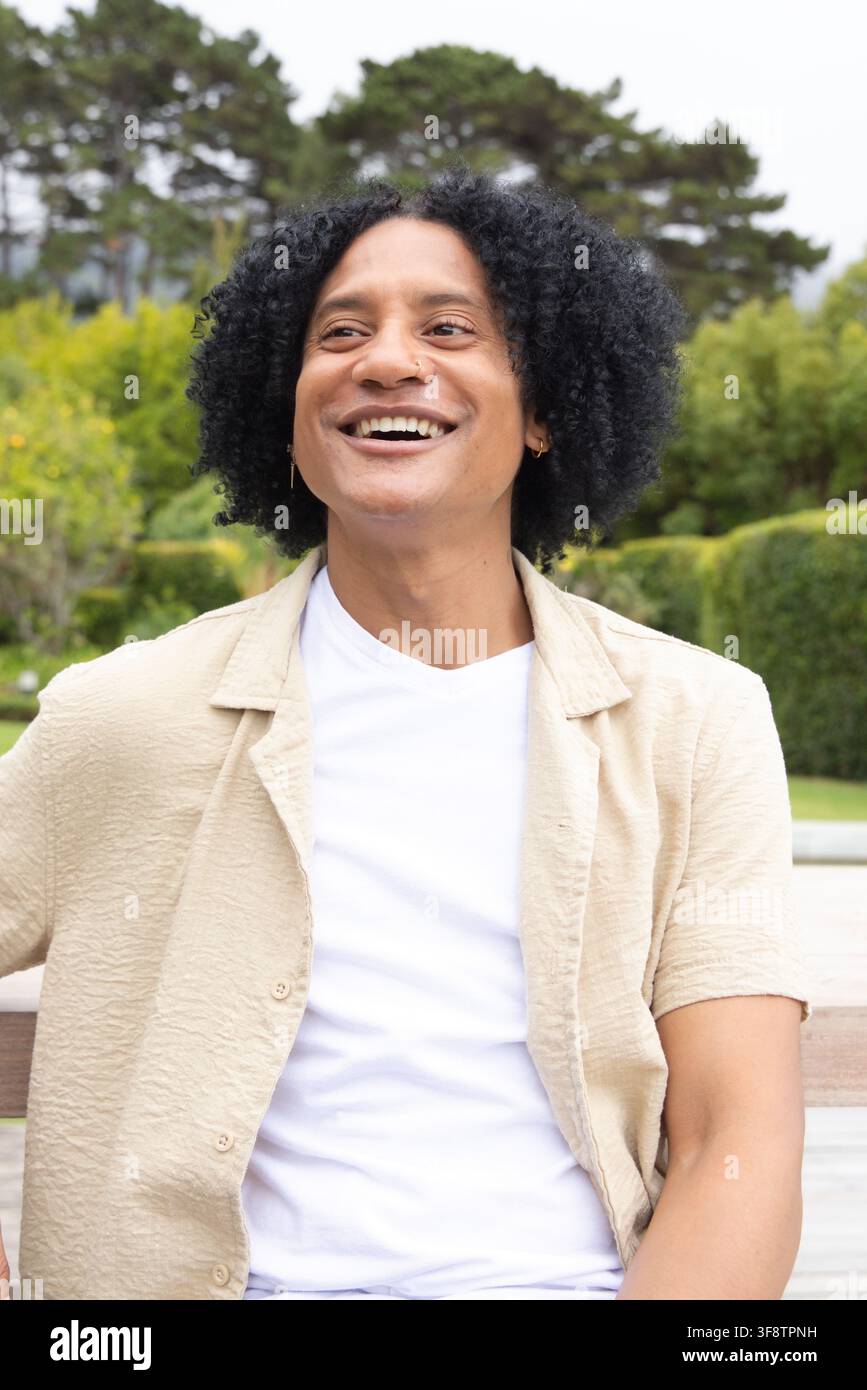 Uomo africano seduto su una panchina di legno nel parco, indossa una camicia beige con un anello al naso, sorride verso l'alto Foto Stock