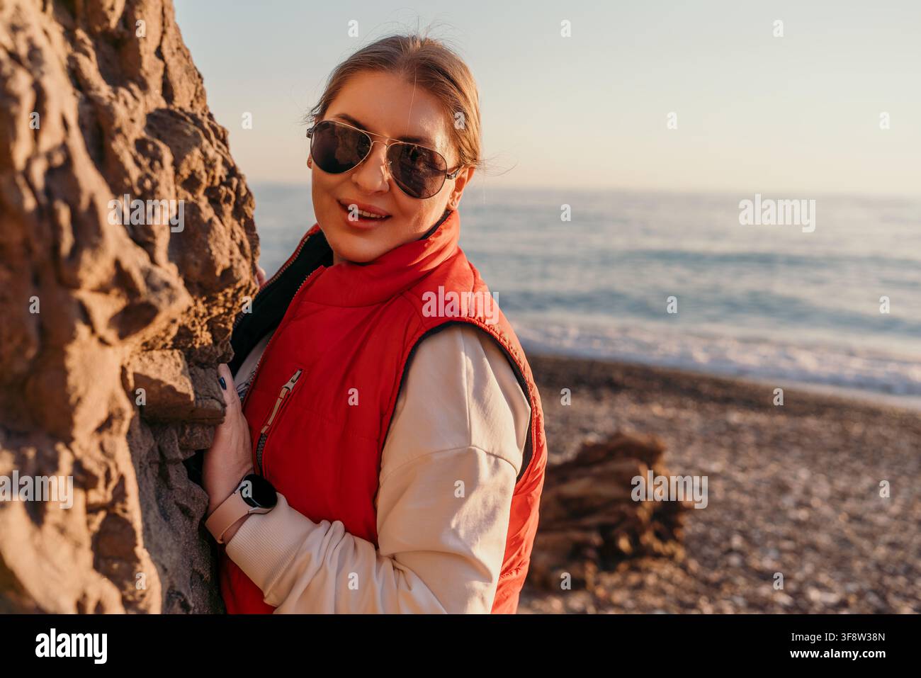 Donna in vacanza al mare, donna sorridente con gilet rosso in piedi vicino a una roccia su una spiaggia di ciottoli al tramonto Foto Stock