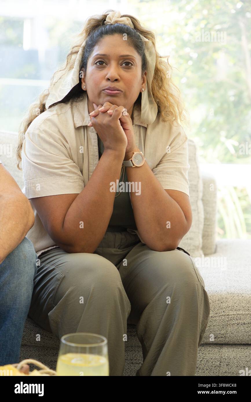 Donna indiana seduta in salotto su un divano chiaro, chinata in avanti con le mani giunte, bicchiere nelle vicinanze Foto Stock