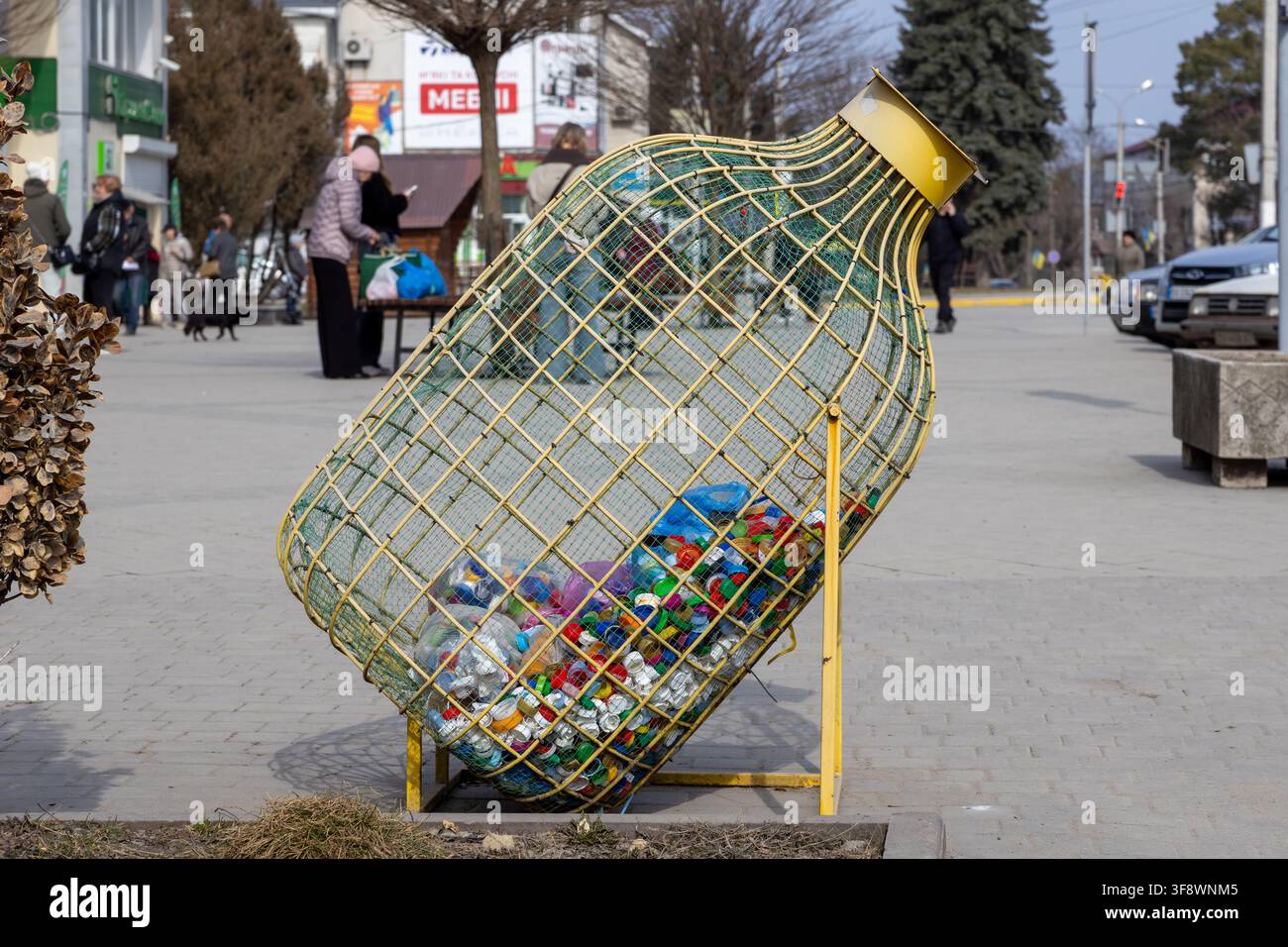 Un contenitore in rete metallica progettato per raccogliere i tappi delle bottiglie di plastica si trova in una piazza pubblica, incoraggiando abitudini ecologiche e la consapevolezza del riciclo con Foto Stock