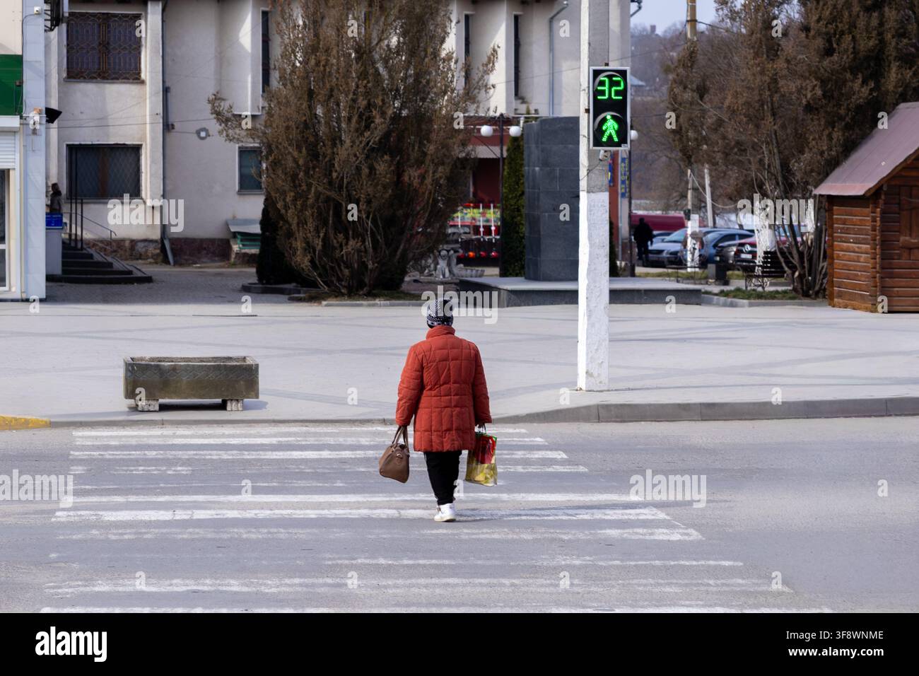 Una persona anziana che porta borse cammina lungo un passaggio pedonale durante il verde, rappresentando la mobilità urbana di routine e la navigazione sicura su strada all'interno di un ambiente tranquillo Foto Stock