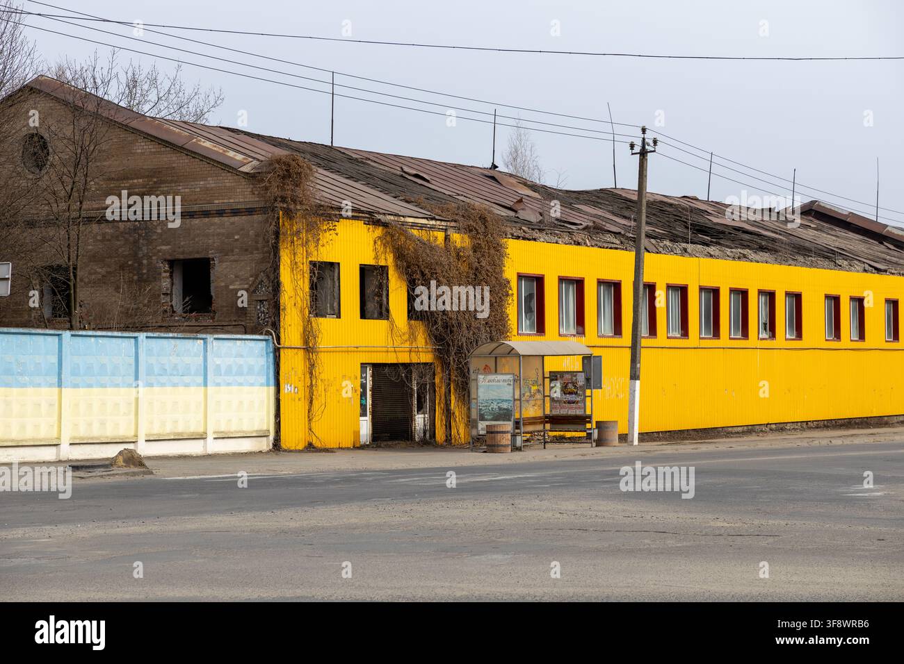 Horodok, Ucraina – 23 marzo 2026: Una struttura industriale abbandonata con il tetto crollato e vite cresciute spontaneamente si trova accanto a una fermata dell'autobus, illustrando l'ambi Foto Stock