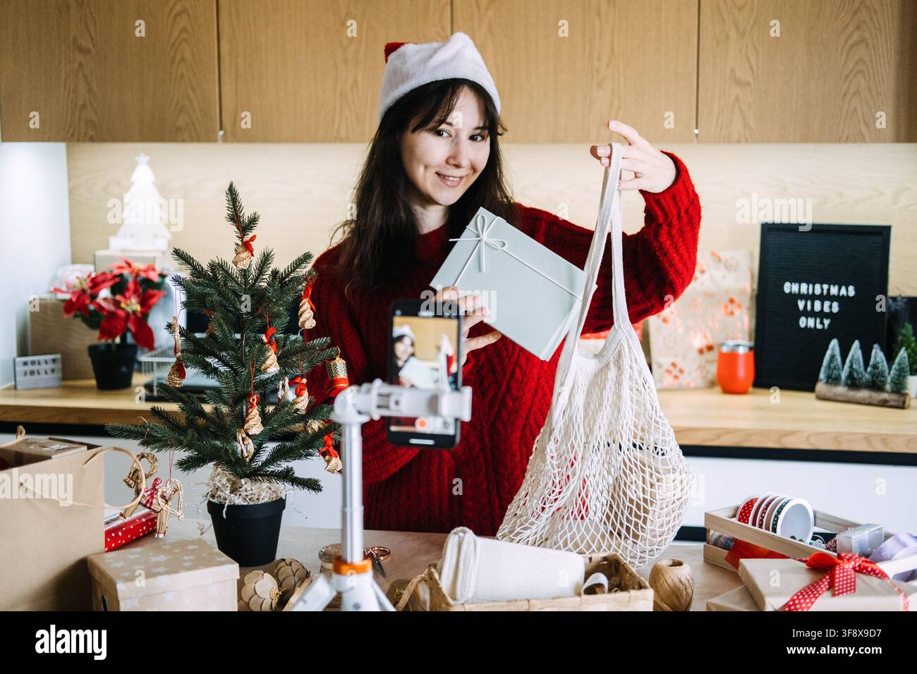 Donna con cappello da Babbo Natale mostra scatola regalo e borsa a rete al telefono su treppiede vicino al cartello CHRISTMAS VIBES ONLY. Recensione di regali di Natale ecologici, regalo sostenibile Foto Stock