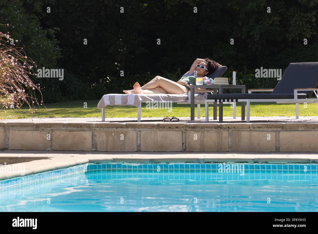 Donna sulla ventina sdraiata su una chaise longue imbottita accanto a una piscina piastrellata, in costume da bagno e occhiali da sole Foto Stock