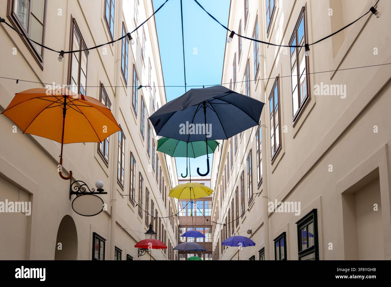 Diversi ombrelli colorati sono sospesi sopra un cortile stretto tra alti edifici chiari, con luci a filo sopra. Foto Stock