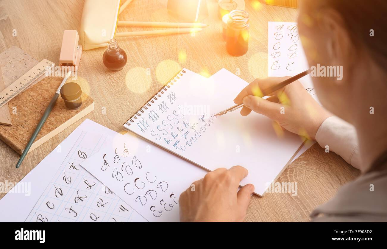 Persona che pratica la calligrafia con una penna a intingimento e inchiostro su un quaderno, circondata da vari utensili per scrivere su una scrivania di legno Foto Stock