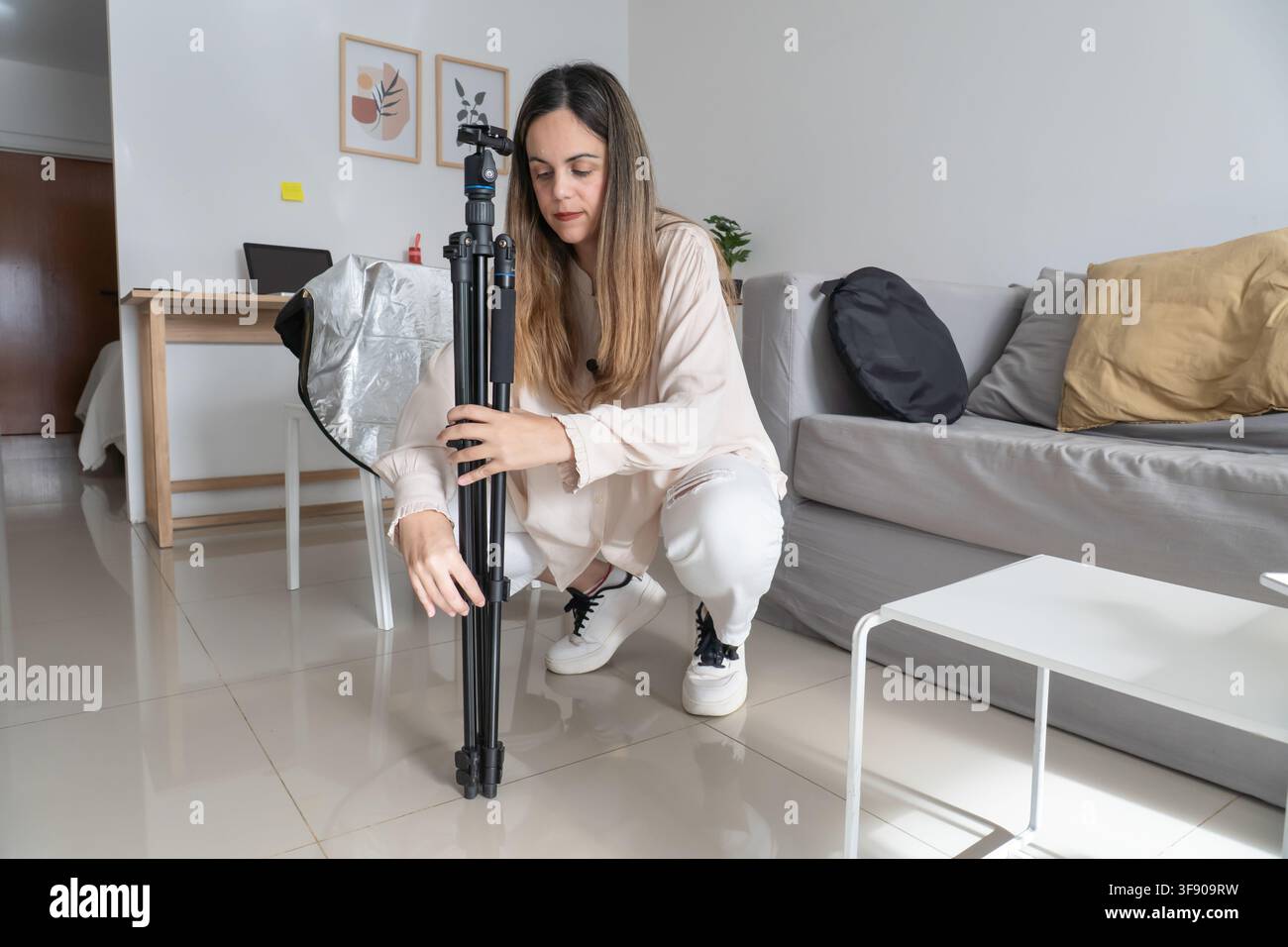 Donna accovacciata per ripiegare le gambe di un treppiede per fotocamera, borsa riflettente appoggiata sul divano, preparazione dell'attrezzatura per il trasporto Foto Stock