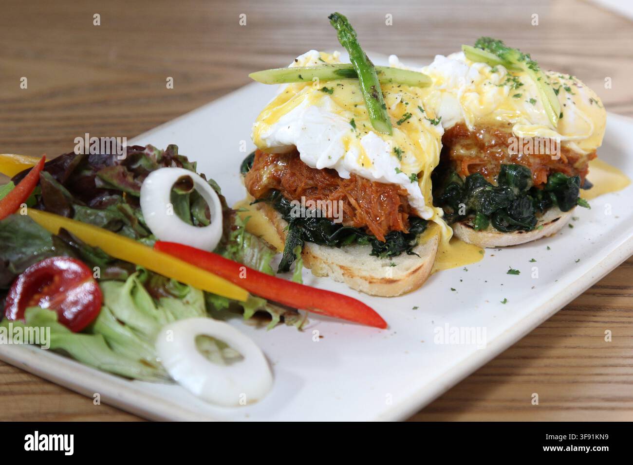 Uova Benedict con pulled pork e spinaci appassiti su muffin inglesi tostati, ricoperti di salsa olandese e asparagi, serviti con una salsa fresca a lato Foto Stock