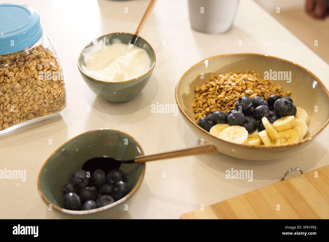 Ciotola di granola su un bancone chiaro con mirtilli, banana, yogurt, barattolo di avena e cucchiai di ottone Foto Stock