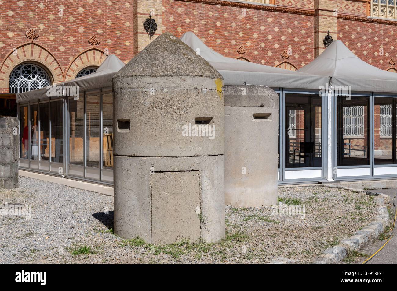 Due caserme cilindriche in cemento con aperture strette si trovano su ghiaia davanti a un edificio storico in mattoni con strutture moderne a baldacchino. Foto Stock