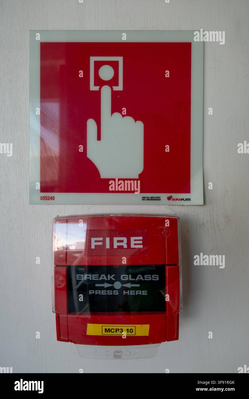 Punto di chiamata allarme antincendio rosso con scatola di vetro infrangibile e segno di pressione manuale montato su un muro bianco, che mostra l'attrezzatura di sicurezza di emergenza all'interno di un edificio. Foto Stock