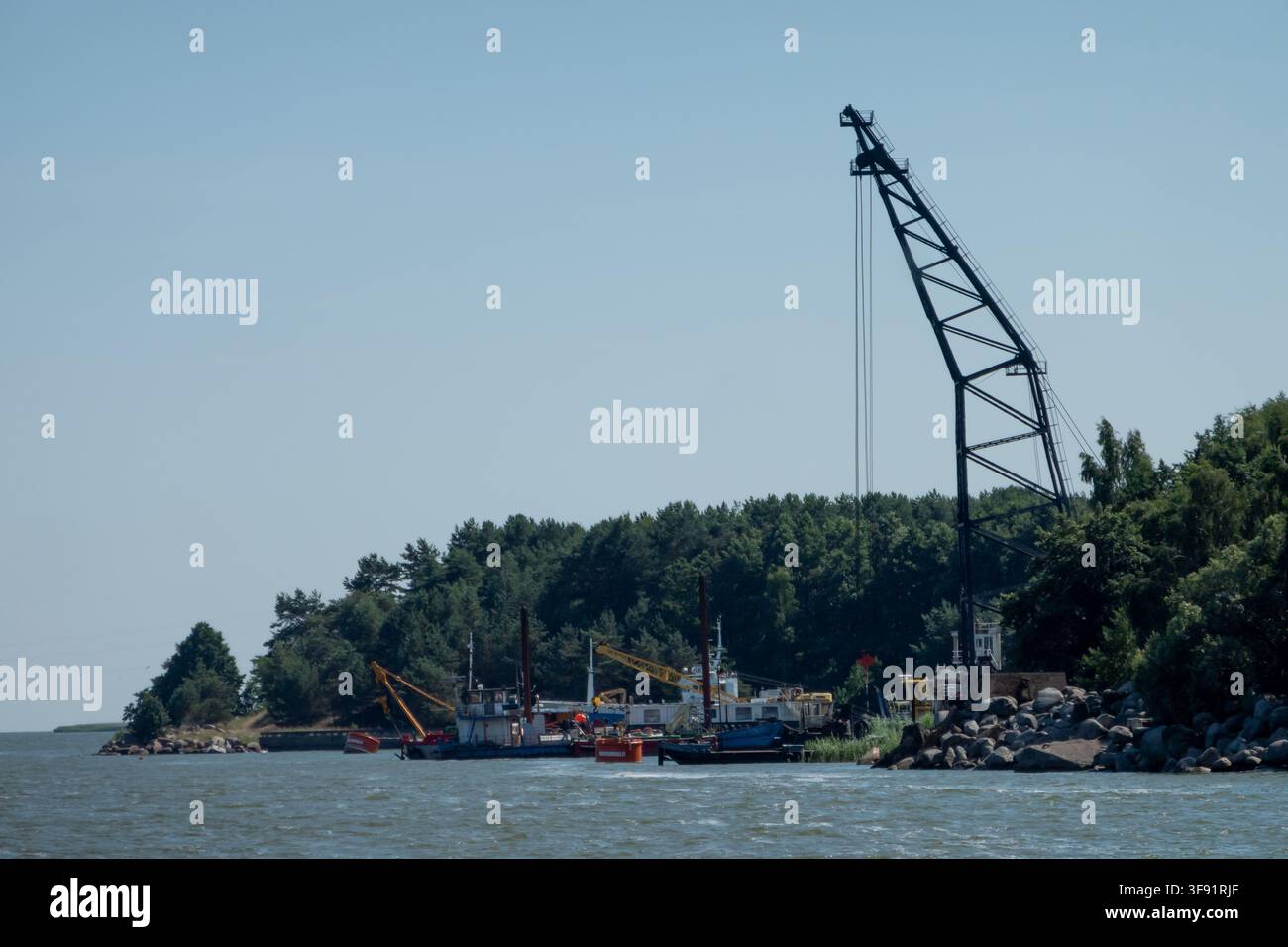 Griglie di costruzione e barche da lavoro vicino a una costa rocciosa e una foresta in una calma giornata costiera, con cielo azzurro e acqua che creano un contrasto industriale. Foto Stock