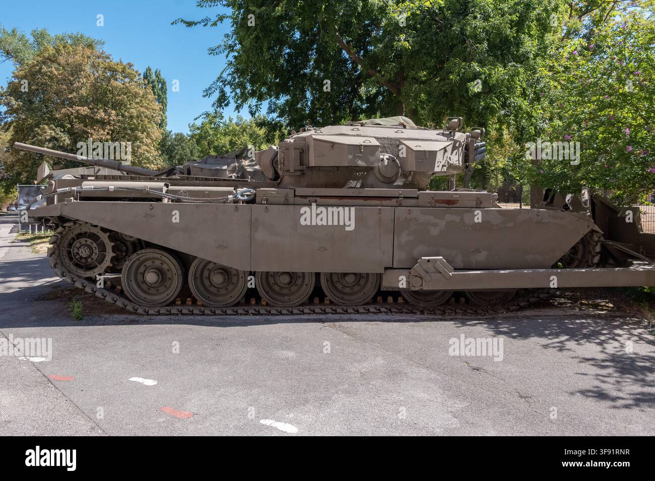 Un carro armato cingolato è mostrato di profilo su un'area pavimentata all'aperto sotto grandi alberi verdi. Foto Stock