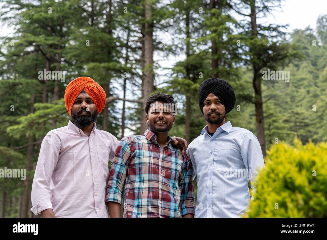 Scatto a bassa angolazione, dal busto in su, di tre amici maschi, tra cui due uomini sikh, in piedi insieme, che guardano l'obiettivo e sorridono mentre posano. Foto Stock