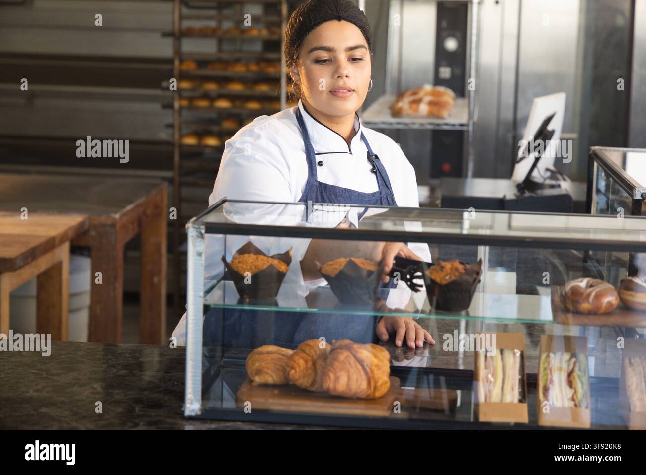 Donna afroamericana in grembiule che prende un muffin con una pinza nella vetrina del bancone della panetteria Foto Stock